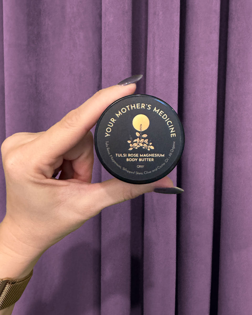A hand with dark nail polish holds a small, round container labeled Tulsi Rose Magnesium Body Butter against deep purple curtains.