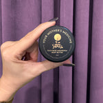 A hand with dark nail polish holds a small, round container labeled Tulsi Rose Magnesium Body Butter against deep purple curtains.