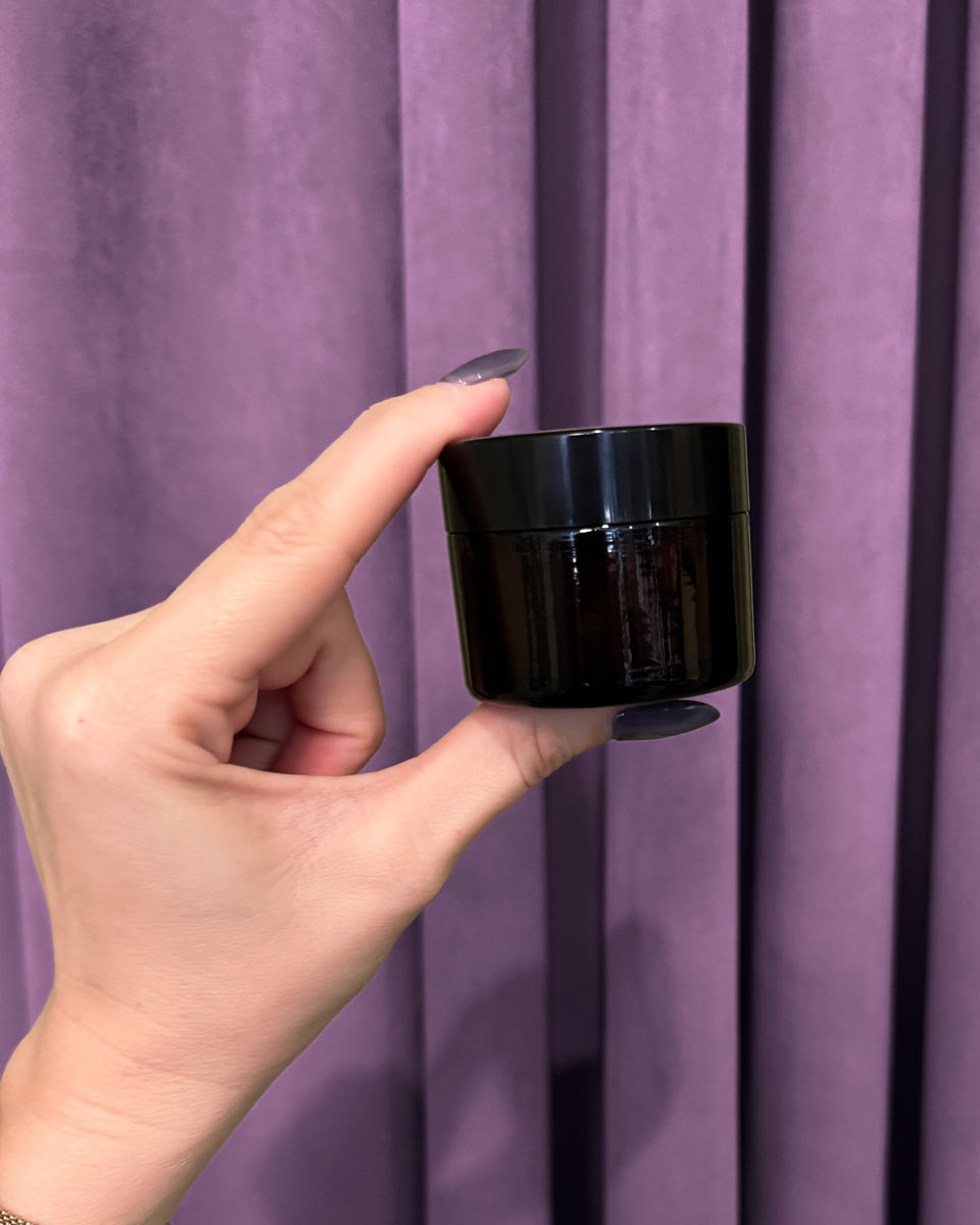 A hand with purple nails holds a small, black, round container of Tulsi Rose Magnesium Body Butter against a purple curtain background.