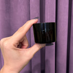 A hand with purple nails holds a small, black, round container of Tulsi Rose Magnesium Body Butter against a purple curtain background.