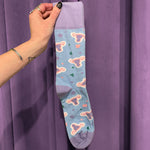 A hand adorned with rings and a bracelet holds the Period Nirvana Cotton Uterus Socks, made from premium combed cotton with pink uterus patterns and colorful shapes, set against a purple curtain background.