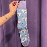 A hand adorned with rings and a bracelet holds the Period Nirvana Cotton Uterus Socks, made from premium combed cotton with pink uterus patterns and colorful shapes, set against a purple curtain background.