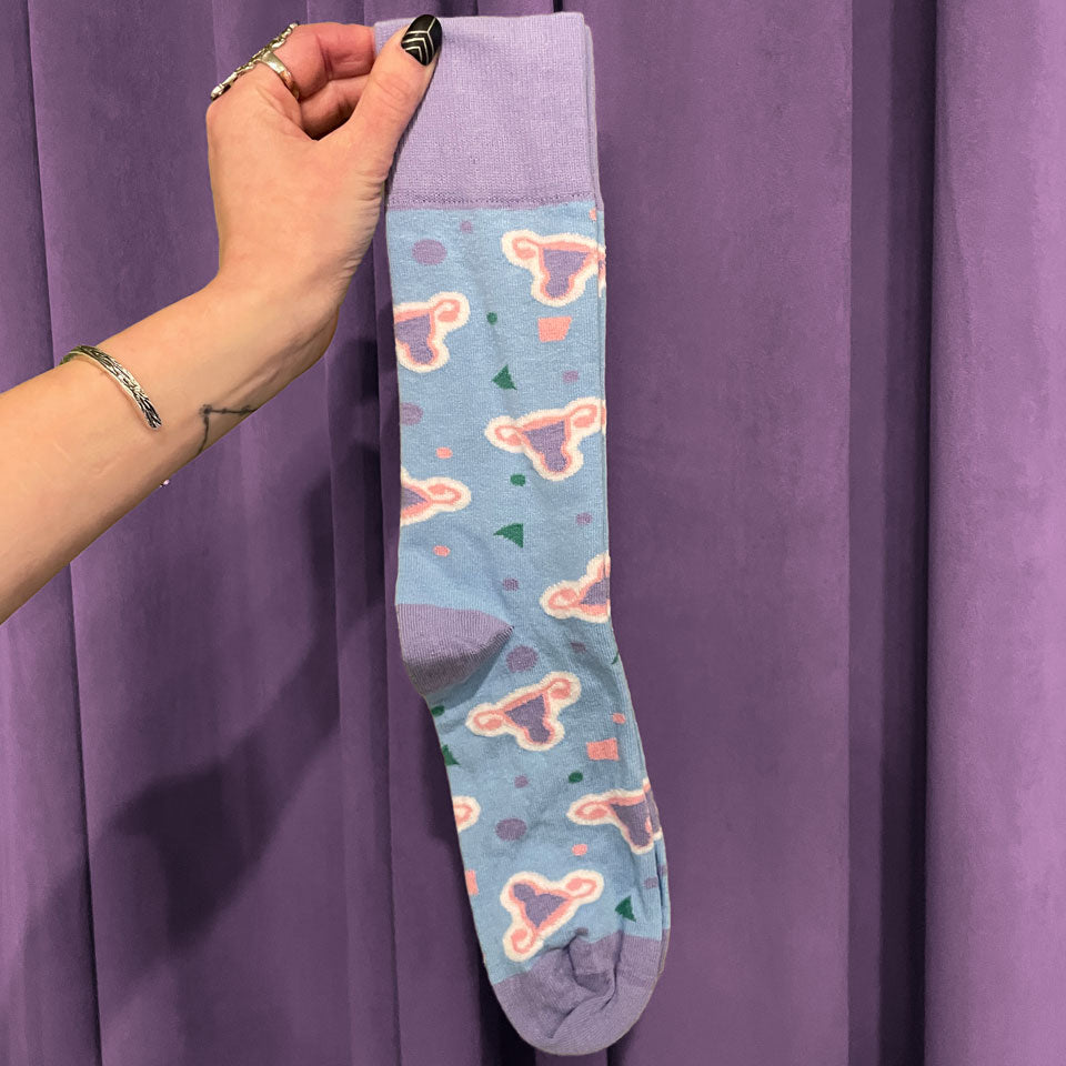 A hand adorned with rings and a bracelet holds the Period Nirvana Cotton Uterus Socks, made from premium combed cotton with pink uterus patterns and colorful shapes, set against a purple curtain background.