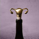 A Uterus Wine Stopper shaped like a uterus and fallopian tubes tops a wine bottle, making a unique reproductive health gift, displayed on a neutral gray background.