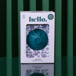 A teal Hello Disc menstrual disc sits on a reflective surface with a green striped background. Its decorative box displays floral designs and highlights leak-proof period protection and a one-size fit.