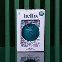 A teal Hello Disc menstrual disc sits on a reflective surface with a green striped background. Its decorative box displays floral designs and highlights leak-proof period protection and a one-size fit.