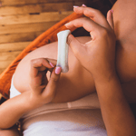 A person with manicured nails holds a white tampon above their lap covered by light shorts on a wooden floor—just as Ohnut Buffer Rings (Set of 4) provide comfortable, body-safe penetration depth control.