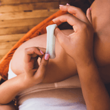 A person with manicured nails holds a white tampon above their lap covered by light shorts on a wooden floor—just as Ohnut Buffer Rings (Set of 4) provide comfortable, body-safe penetration depth control.