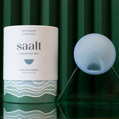 A Saalt Disc and its packaging are shown on a green, textured background. The cylindrical box labeled "Saalt Disc" is next to the blue disc on a clear stand, highlighting easy removal and comfortable size options.
